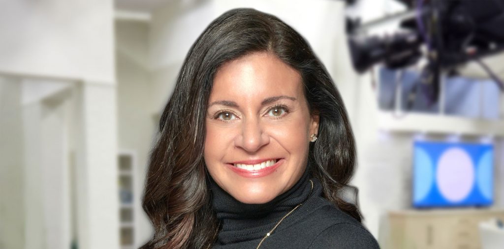 Stacy Bowe in a dark grew turtleneck and gold chain necklace, standing in a studio.